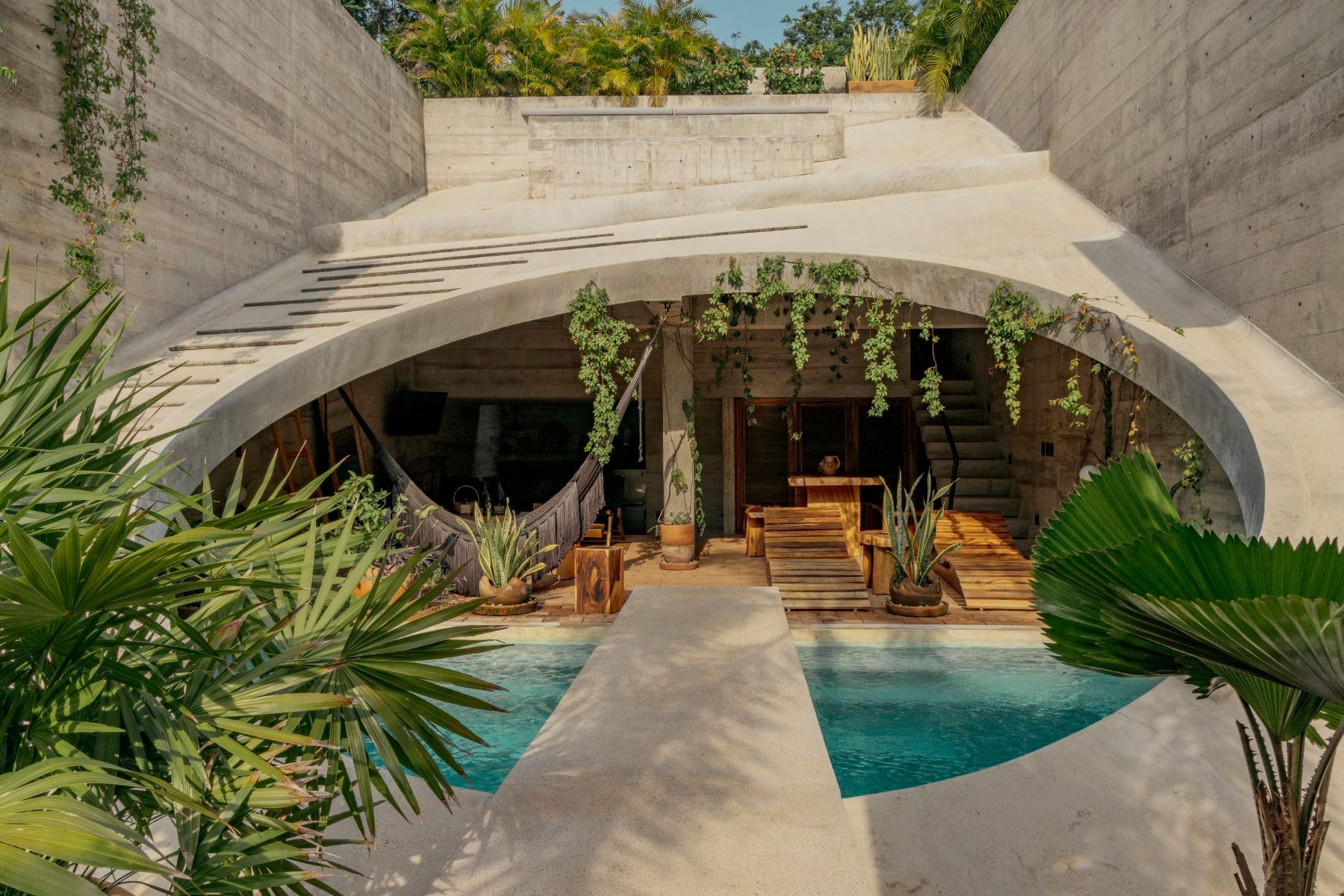 The oval concrete opening of Casa Hezbo, with the pool, a concrete bridge and the open living area under hanging plants